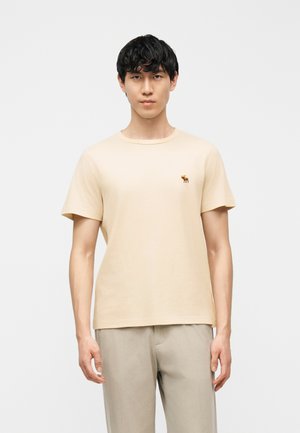 Jonge man met kort krullend zwart haar, gekleed in een effen beige T-shirt met een klein elandlogo op de borst en beige broek.