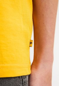 Camiseta de algodón amarilla con etiqueta en la costura lateral; presenta una textura suave, mangas cortas y un corte recto en el dobladillo.