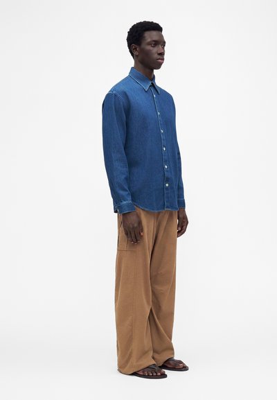 Homme debout portant une chemise en jean bleue à manches longues, un pantalon marron ample et des sandales noires sur un fond blanc uni.