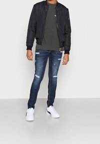 Chaqueta bomber negra, camiseta gris, vaqueros pitillo azules desgastados con parches claros y zapatillas blancas. Conjunto sencillo y casual.