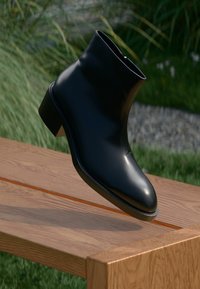 Black leather ankle boot with a smooth finish, rounded toe, and a low block heel, displayed on a wooden surface.