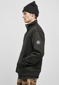 Juoda zip-up striukė su aukštu apykakliu, megztu paviršiumi, šoninėmis kišenėmis ir rankovės ženkleliu. Dėvima su juodu kepurėle ir kamufliažo kelnėmis.