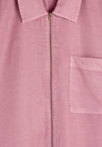 Różowa bawełniana koszula z metalowym zamkiem błyskawicznym na środku oraz prostokątną kieszonką na piersi po prawej stronie.