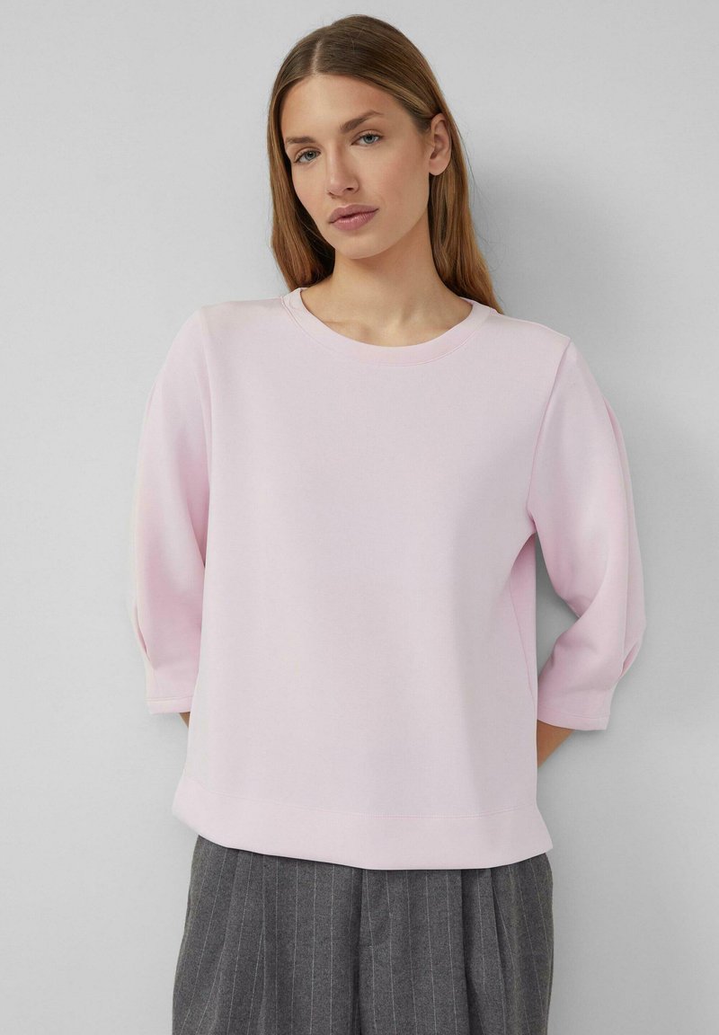 Hellrosa Sweatshirt mit dreiviertel Ärmeln, rundem Ausschnitt und lockerer Passform. Kombiniert mit grauen, gestreiften Hosen. Glatte Textur.