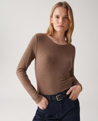 Young woman wearing a brown ribbed long-sleeve top tucked into dark blue jeans with a black belt, hands in pockets, neutral background.