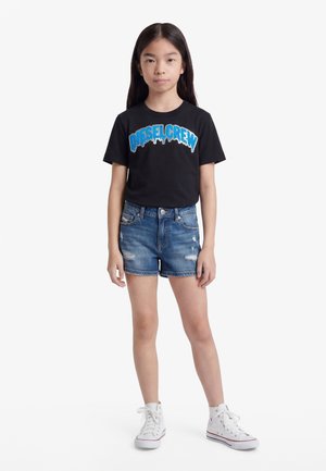 Ung jente står iført svart "DIESEL CREW" t-skjorte, blå denimshorts og hvite høye sneakers på hvit bakgrunn.