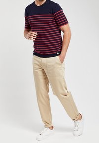 Hombre que lleva una camiseta de rayas en azul marino y rojo, pantalones beige con bolsillos de parche y zapatillas blancas, de pie con una mano en el bolsillo.