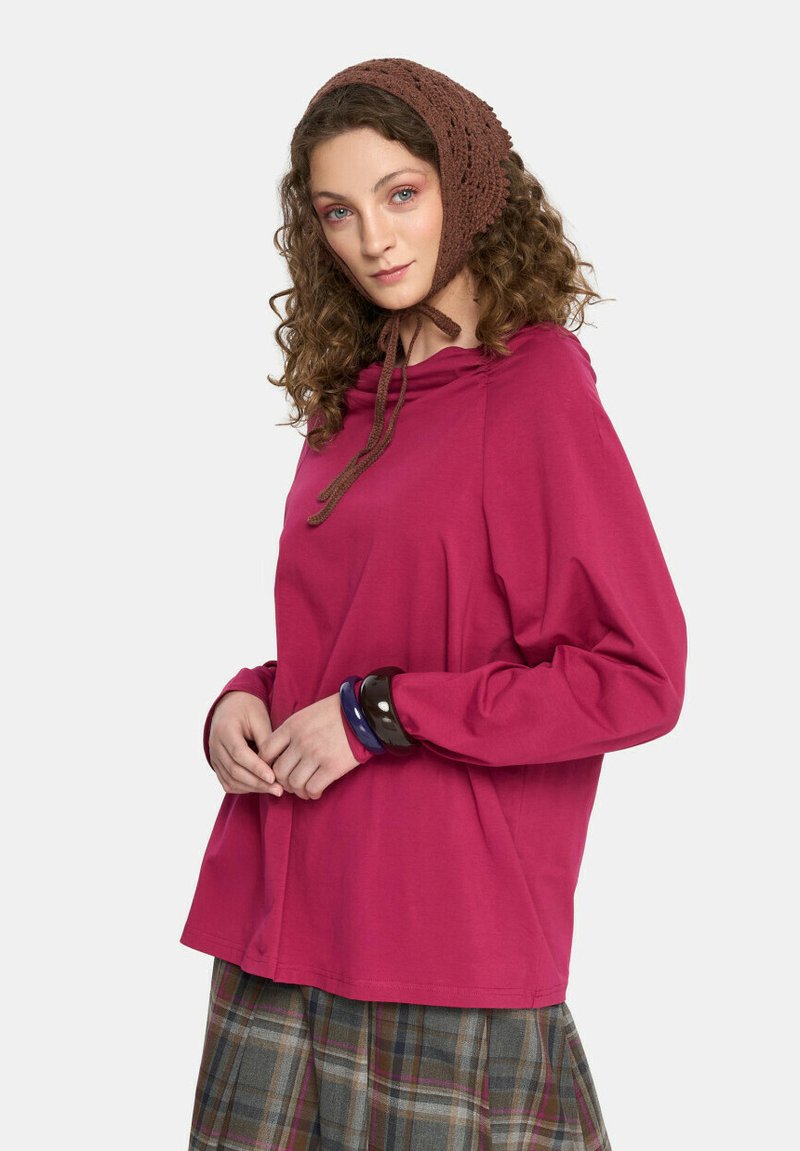 Fioletowa bluzka z długim rękawem i marszczonym dekoltem oraz sznurkiem, zestawiona z spódnicą w kratę. Dodatki to naszyjniki w stylu warstwowym i brązowy dziany chustka na głowę.