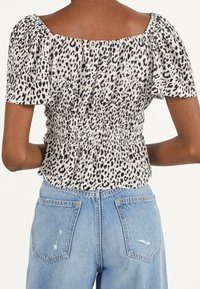 Widok z tyłu osoby noszącej czarną i białą bluzkę w panterkę z krótkim rękawem, z marszczonym pasem, w połączeniu z jasnoniebieskimi dżinsami z wysokim stanem.