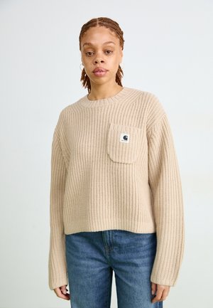 Maglione lavorato a maglia beige con collo rotondo, texture a coste e tasca davanti. Abbinato a jeans in denim blu. Logo del marchio visibile sulla tasca.