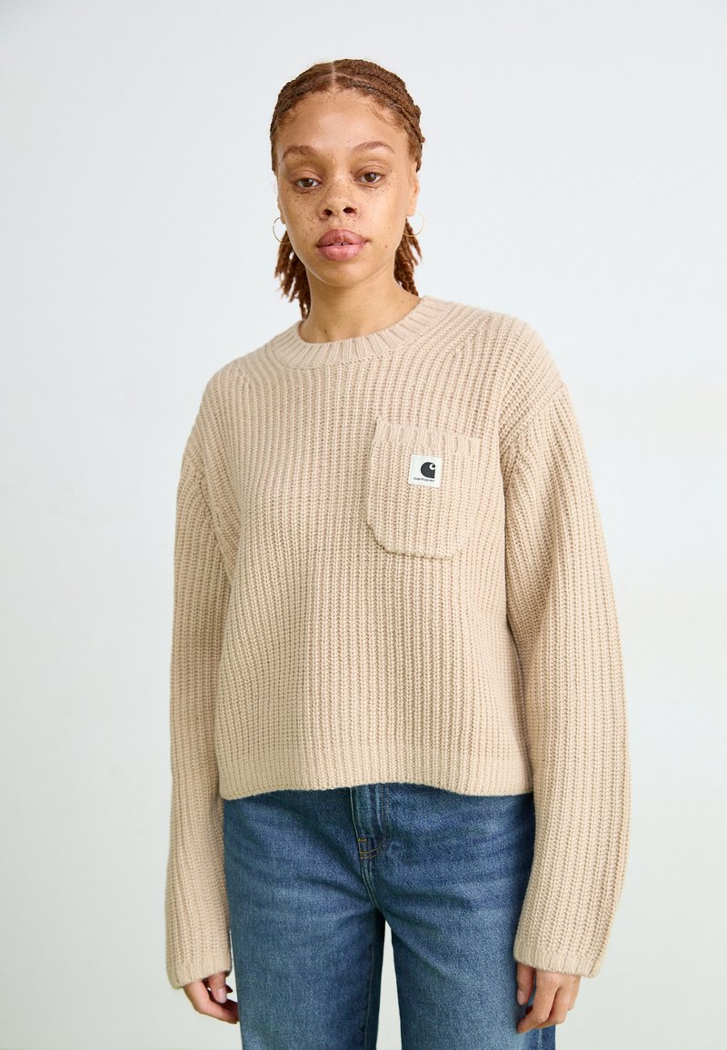 Maglione lavorato a maglia beige con collo rotondo, texture a coste e tasca davanti. Abbinato a jeans in denim blu. Logo del marchio visibile sulla tasca.