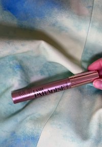 Tusz do rzęs Lash Paradise w metaliczno-różowej tubie z czarnym tekstem na miękkim, teksturowanym materiale w stonowanych odcieniach zieleni i niebieskiego.