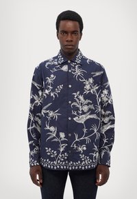 Tummansininen paita, jossa on valkoista kukka- ja lintikirjontaa, napitus edessä, pitkät hihat ja terävä kaulus. Silkkinen kangas ja rento malli.