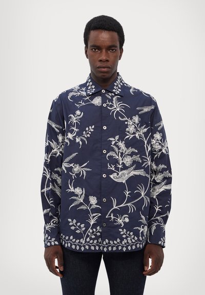 Camisa azul marino con bordado blanco de flores y aves, con botones en la parte delantera, mangas largas y cuello puntiagudo. Tejido suave y corte relajado.