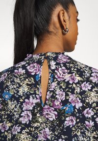 Femme avec queue de cheval foncée portant une boucle d'oreille anneau en argent et un chemisier bleu marine à fleurs avec fermeture au dos en forme de trou de serrure et des fleurs violettes, bleues et crème.