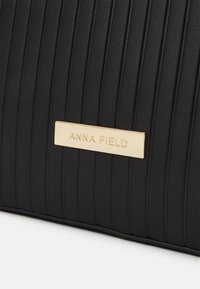 Black handbag featuring vertical ribbed texture, metallic gold brand plate "ANNA FIELD," and a smooth faux leather finish.