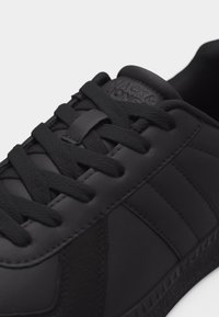 Nahaufnahme eines schwarzen Ledersneakers mit schwarzen Schnürsenkeln, gepolstertem Schaft und eingeprägtem Logo auf der Zunge.