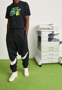 Czarna koszulka Nike z kolorowym logo, czarne dresy z białymi akcentami i kremowe trampki. W tle znajduje się ksero na zielonym dywanie.