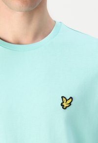 T-shirt en coton bleu clair avec un col rond, en tissu texturé, présentant un petit logo d'aigle noir et jaune sur la poitrine gauche.