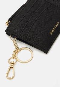 Zwarte gestructureerde portefeuille met gouden hardware, voorzien van een sleutelhanger en sluiting, gebosseerd met "ANNA FIELD" en gedetailleerde stiksels.