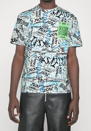 Person wearing a white short-sleeve T-shirt with mixed black and blue "Just Cavalli" text patterns and a green pocket patch, paired with black leather pants.