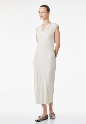 Vrouw staat in een mouwloze, gebroken witte, geplooide midi-jurk met een V-hals, gecombineerd met slangenprint platte schoenen en gouden oorringen.