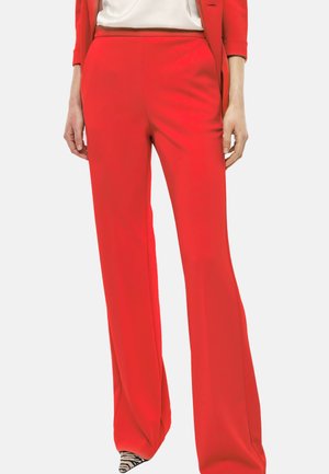 Donna che indossa pantaloni ampi rosso vivo, un blazer coordinato e scarpe a punta con stampa zebra, in piedi con le mani rilassate ai lati.