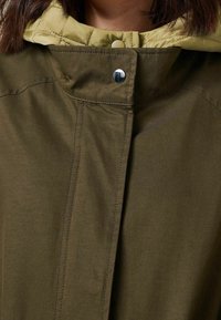 Veste vert olive avec une capuche matelassée vert clair, dotée d'une fermeture à pression et de détails de couture visibles sur le tissu.