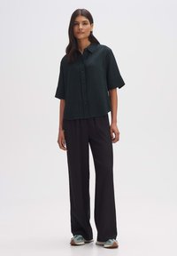 Dark green, textured short-sleeve shirt with button front, paired with loose black pants and light-colored sneakers. Simple, modern design.