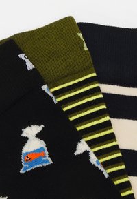 Tre par sokker: ét par sort med fiskedesign, ét par olivengrønne med vandrette striber og ét par marineblå med kremfarvede striber. Alle er strikket stof.