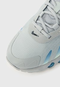 Adidaș sportiv gri deschis și albastru, cu partea superioară din plasă, mic logo Nike negru lângă șireturi și talpă de cauciuc, vedere înclintă a zonei degetelor.