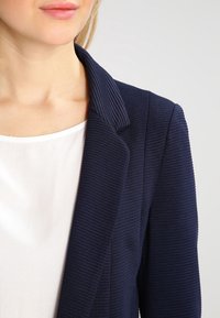 Námořnický žebrovaný blazér se strukturovaným klopem, v kombinaci s bílou blůzou. Tkanina má texturovaný povrch pro přidaný detail.