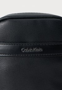 Saco de couro preto com superfície texturizada, design elegante e logotipo "Calvin Klein" em prata embossado numa alça horizontal.