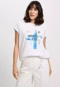 Biała koszulka z krótkim rękawem z niebieskimi i szarymi grafikami w stylu pociągnięcia pędzlem. Czarny napis brzmi: "APPRÉCIE CHAQUE INSTANT DE LA VIE."