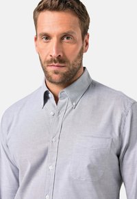 Vaaleanharmaa nappipaito, jossa on kaulus ja pitkät hihat, yhdellä rinnan taskulla ja hienovaraisella tekstuurilla. Ominaisuuksiin kuuluvat valkoiset napit ja ommellut yksityiskohdat.