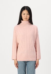 Svetra svetlo roza turtleneck z teksturiranim vzorcem in rebrastimi manšetami. Ohlapen kroj z dolgimi rokavi, oblečen čez svetlo modre kavbojke.