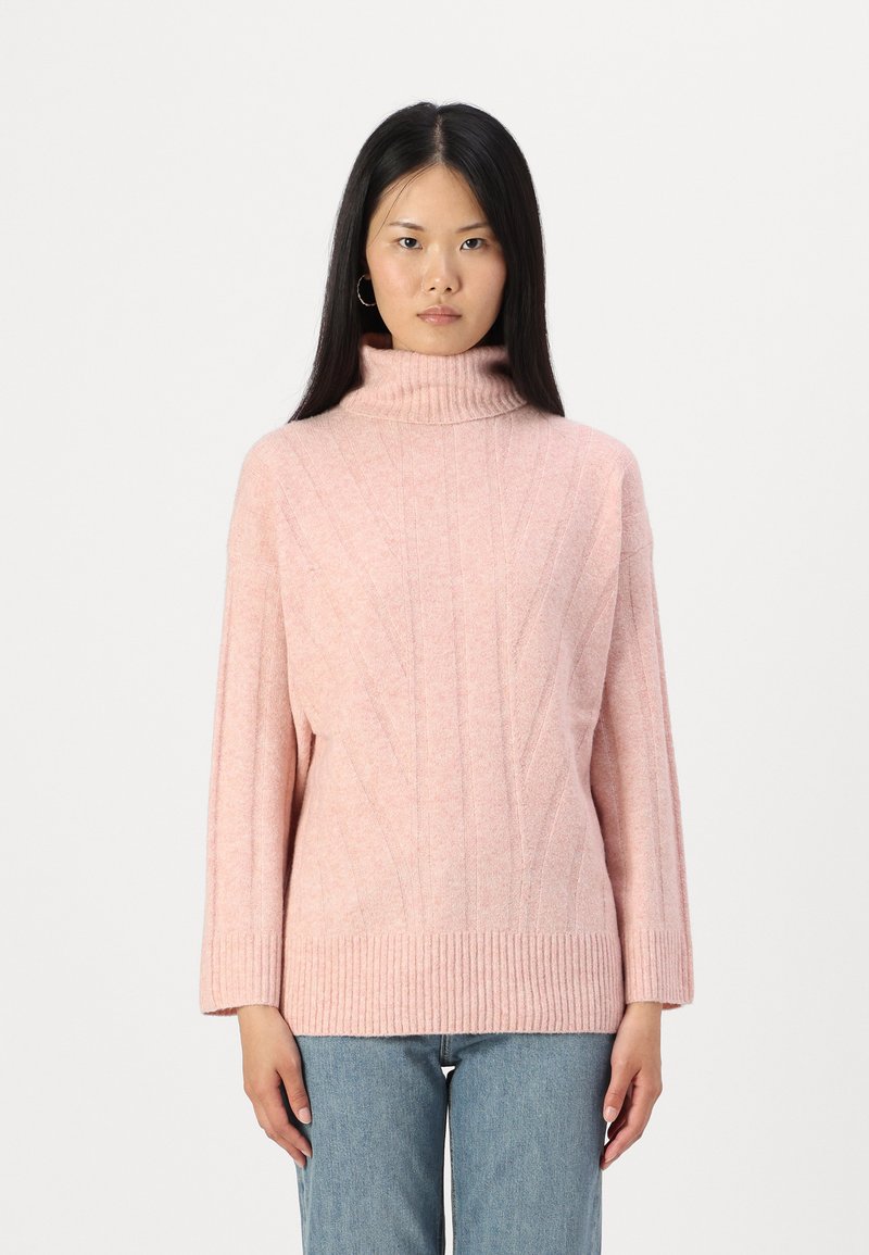 Svetra svetlo roza turtleneck z teksturiranim vzorcem in rebrastimi manšetami. Ohlapen kroj z dolgimi rokavi, oblečen čez svetlo modre kavbojke.