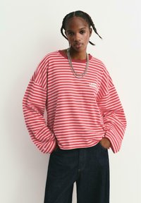 Giovane con capelli intrecciati, che indossa una maglietta a maniche lunghe rossa e rosa a righe, catene d'argento e jeans scuri, in piedi con le mani in tasca.