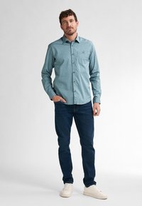 Camicia azzurra chiara con un sottile motivo a pois scuri, maniche lunghe e colletto, abbinata a jeans blu scuro e scarpe da ginnastica bianche.
