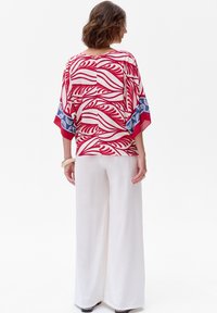 Rood en wit gebloemd shirt met vloeiende mouwen, gecombineerd met brede witte broek. Het shirt heeft een botanisch ontwerp en blauwe accenten.