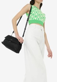 Borsa nera strutturata con chiusura a patta, hardware argentato e tracolla regolabile, abbinata a un top corto a motivi verdi e pantaloni bianchi a gamba larga.
