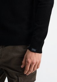 Schwarzer Strickpullover mit gerippten Bündchen, ausgestattet mit einem Logobadge am Ärmel. Kombiniert mit braunen, strukturierten Cargo-Hosen.