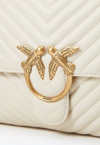 Bežā krāsā izšūta ādas rokassoma ar zelta toņa gredzenu aizdari un diviem putnu akcentiem. Ievērojama chevron šuvju detaļa visā somas virsmā.