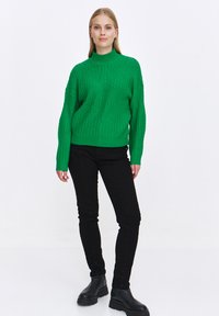 Przesadzony zielony sweter dziany z wysokim kołnierzem, z teksturowanymi wzorami. W zestawie z czarnymi spodniami slim fit i masywnymi czarnymi botkami.