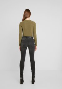 Jeans skinny pretos com cintura alta, combinados com uma blusa de mangas longas em verde-oliva. O modelo usa botins pretos de salto grosso.