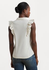 Blouse sans manches beige clair avec des accents de volants aux épaules, un col, et un tissu doux et texturé. Portée avec un jean en denim foncé.