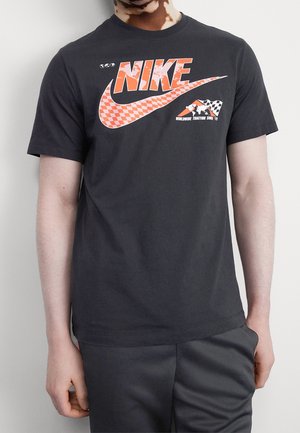 Person trägt schwarzes Nike-T-Shirt mit orange-kariertem Swoosh und Text „Worldwide traction since '72“ vor weißem Hintergrund.