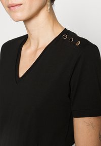 Schwarzes V-Ausschnitt-T-Shirt mit kurzen Ärmeln, das drei goldfarbene dekorative Knöpfe auf der Schulter aufweist. Glatte Stofftextur.