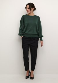 Maglione verde con paillettes, dall'aspetto morbido, maniche lunghe e orlo a costine, abbinato a pantaloni neri su misura e décolleté neri a punta.