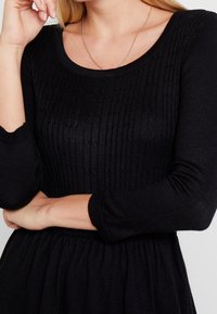 Robe en maille noire avec une texture côtelée, manches longues et encolure ronde ; présente une taille froncée et un tissu doux.